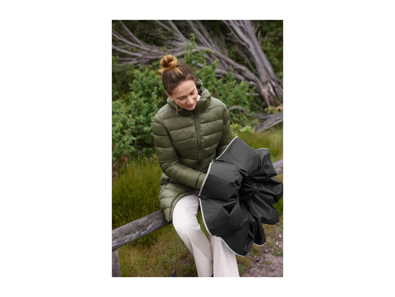 A woman in a green puffer jacket and white pants sits on a wooden fence, holding a black umbrella.