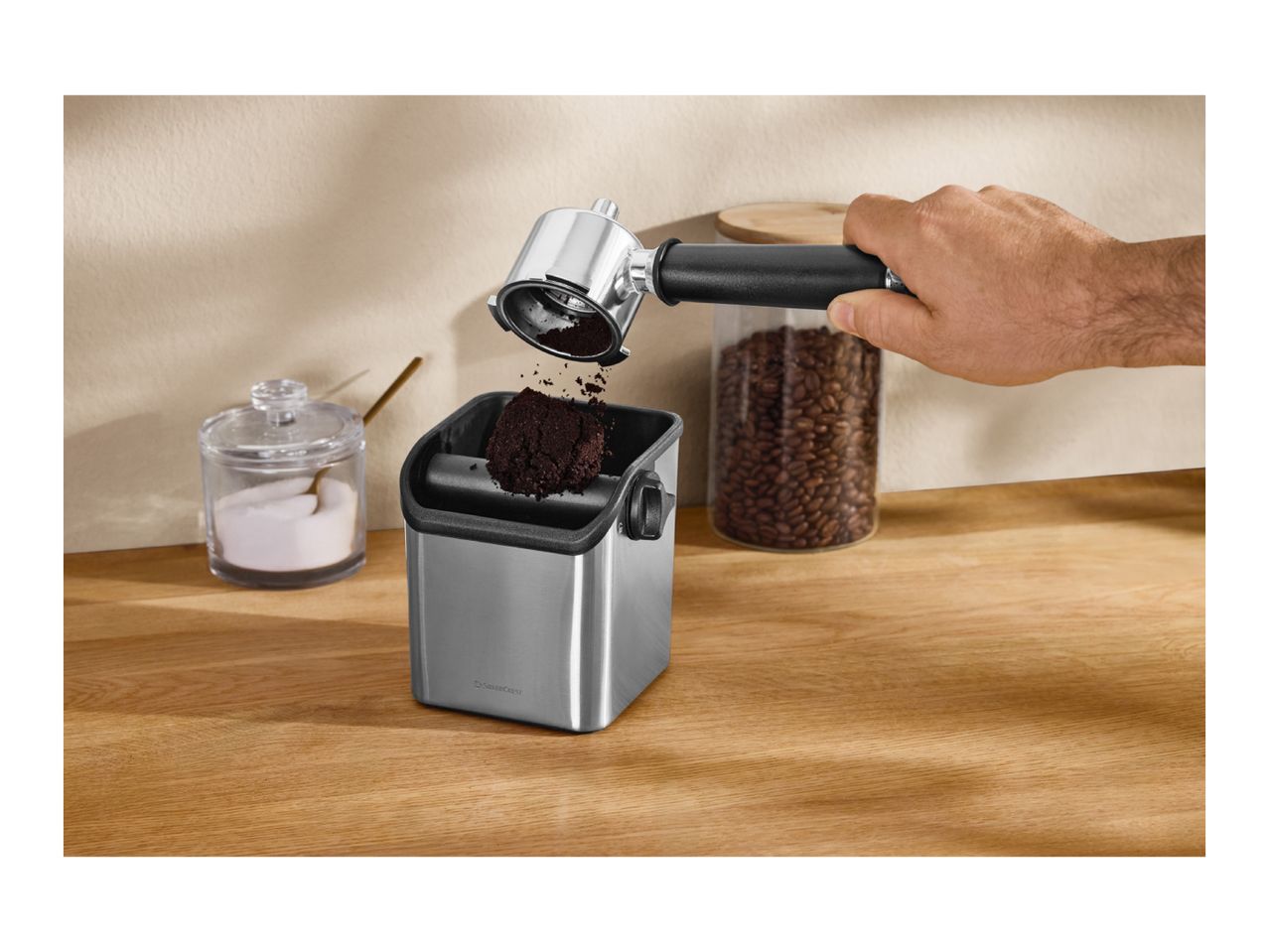 A hand knocking a portafilter with used coffee grounds into a stainless steel knock box.