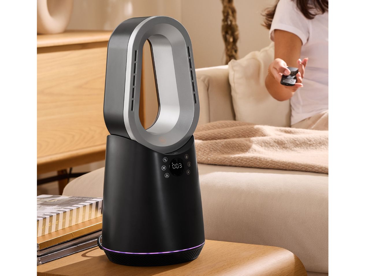 Black bladeless fan with a digital display on a wooden table, with a person holding a remote in the background.