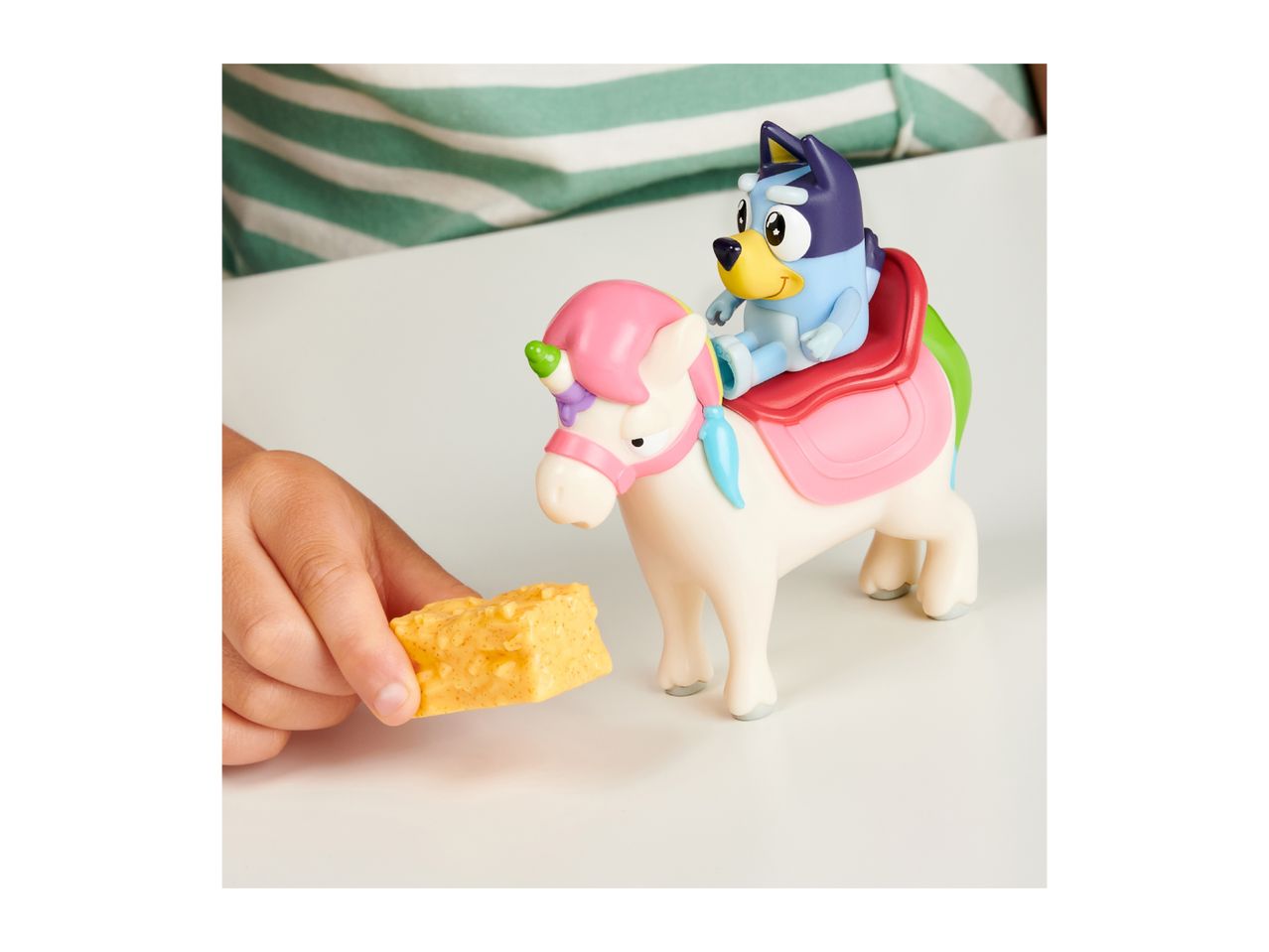 Child's hand holding a toy cheese block, next to a Bluey figure riding a unicorn toy.