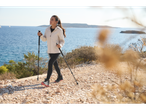 Mulher de casaco bege e leggings de corrida pretas, caminhando com bastões junto ao mar.