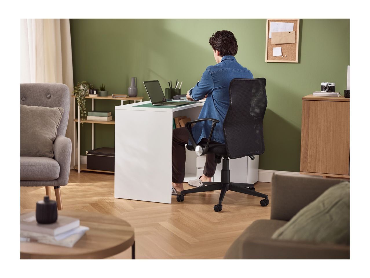 Man working on a laptop at a white desk in a home office with green walls.