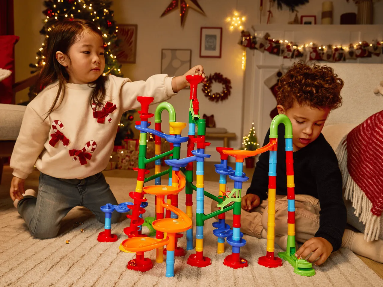 Zwei Kinder spielen mit einer bunten Murmelbahn vor einem Weihnachtsbaum.