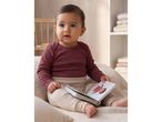 Baby in a long-sleeved bodysuit and leggings, holding a board book with an illustration of a person.