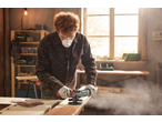 Man sanding wood with an orbital sander in a workshop, wearing safety glasses and a mask.