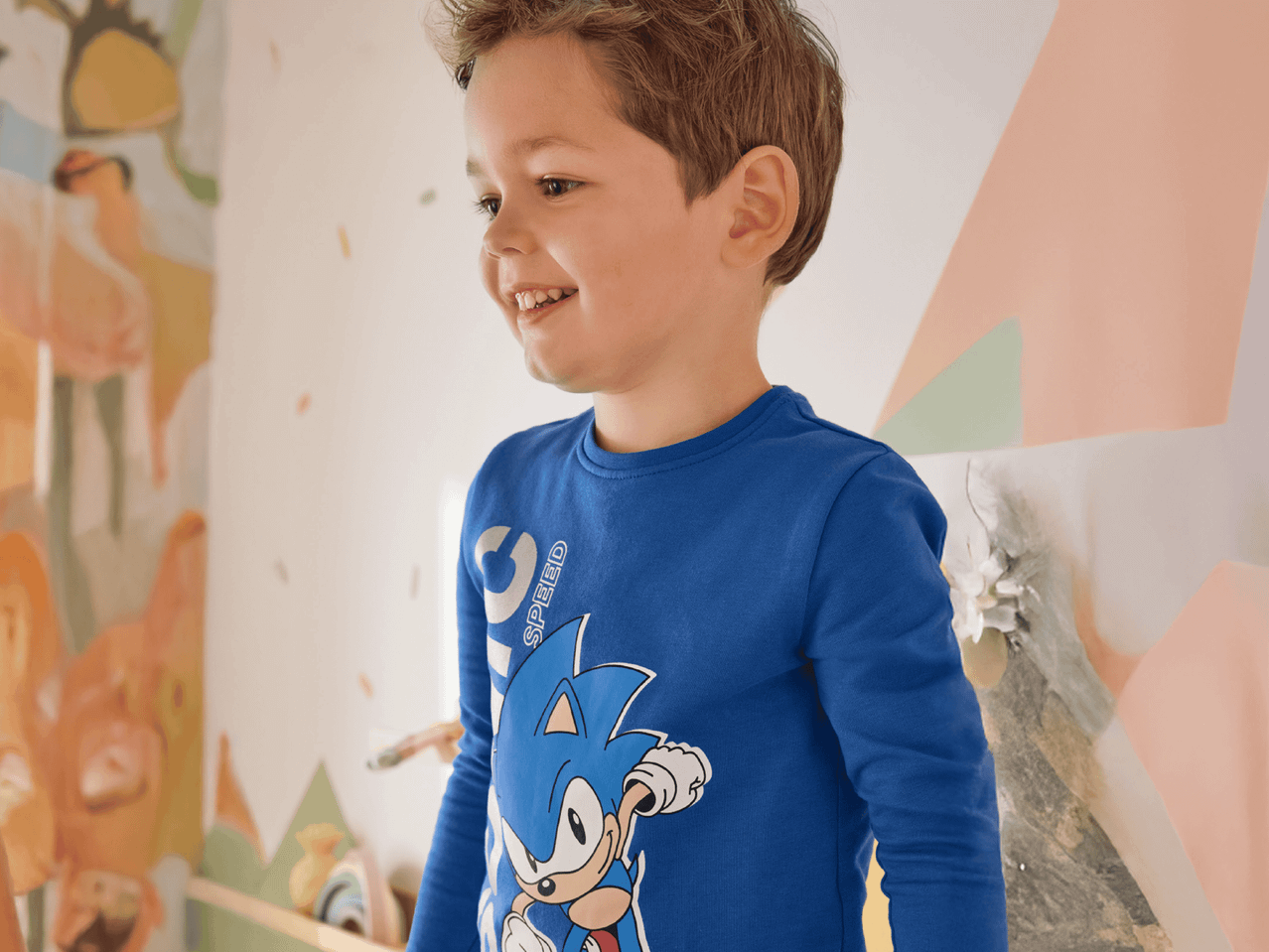Junge in blauem Langarmshirt mit Sonic-Aufdruck, lachend vor einer bunten Wand.