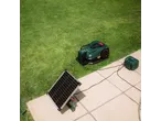 Robotic lawnmower charging via a solar panel and power station on a patio next to grass.