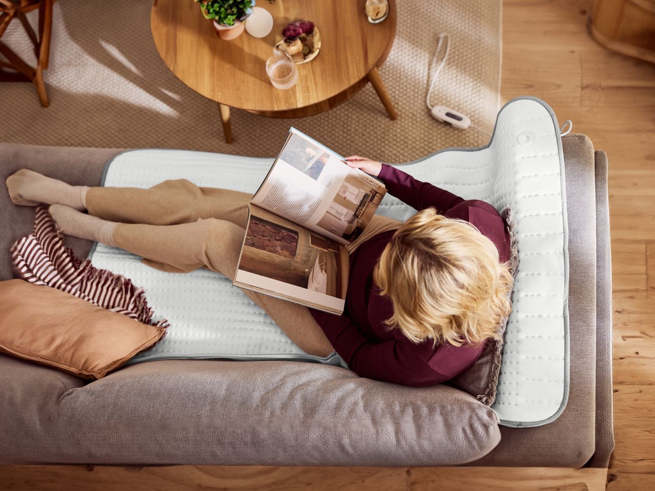 Woman relaxing on a sofa with a heated blanket, reading a magazine.