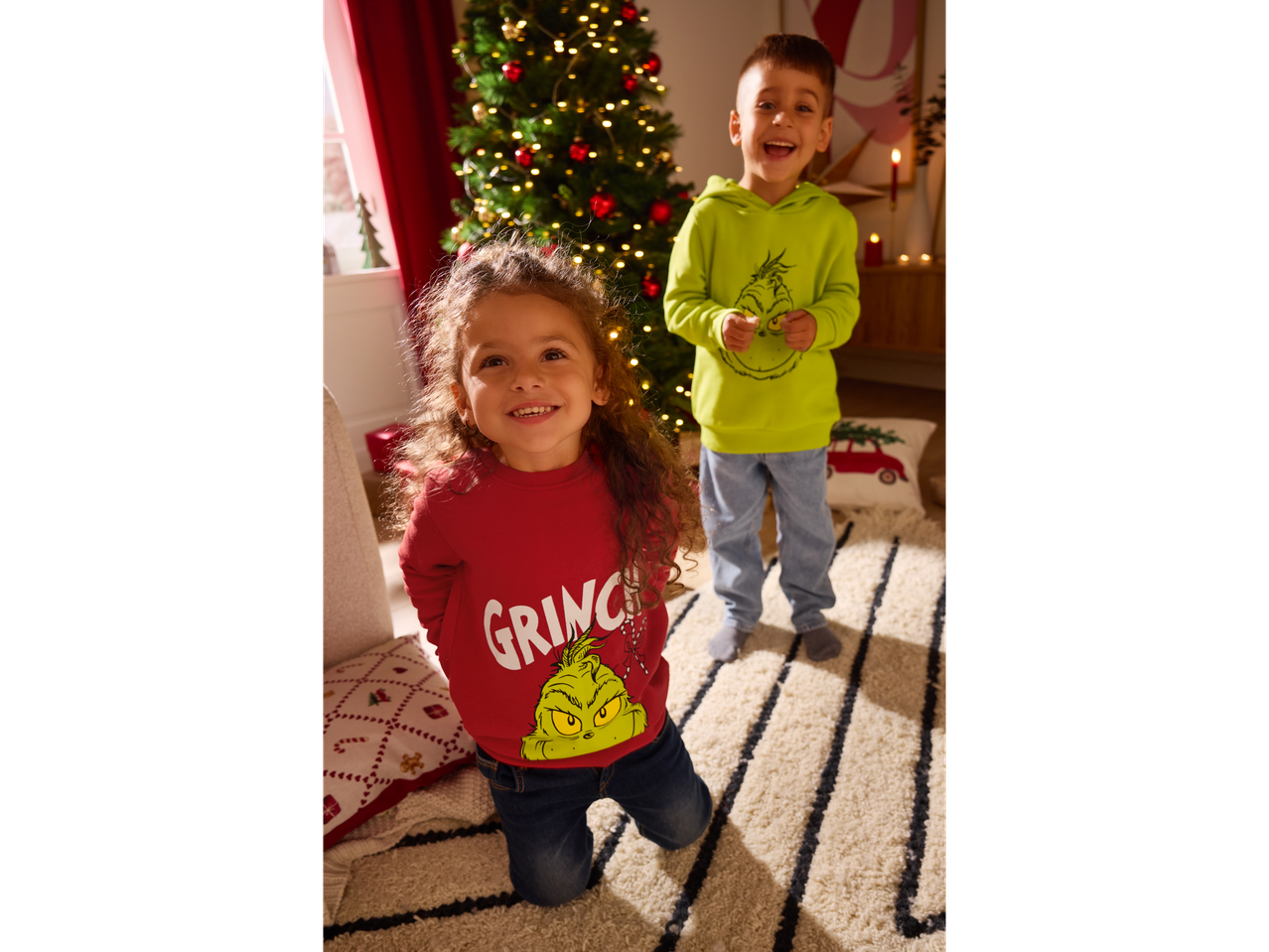 Du vaikai su Grinčo džemperiais priešais Kalėdų eglutę.