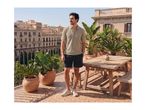 Man in a polo shirt and shorts on a sunny balcony with potted plants and a wooden table.