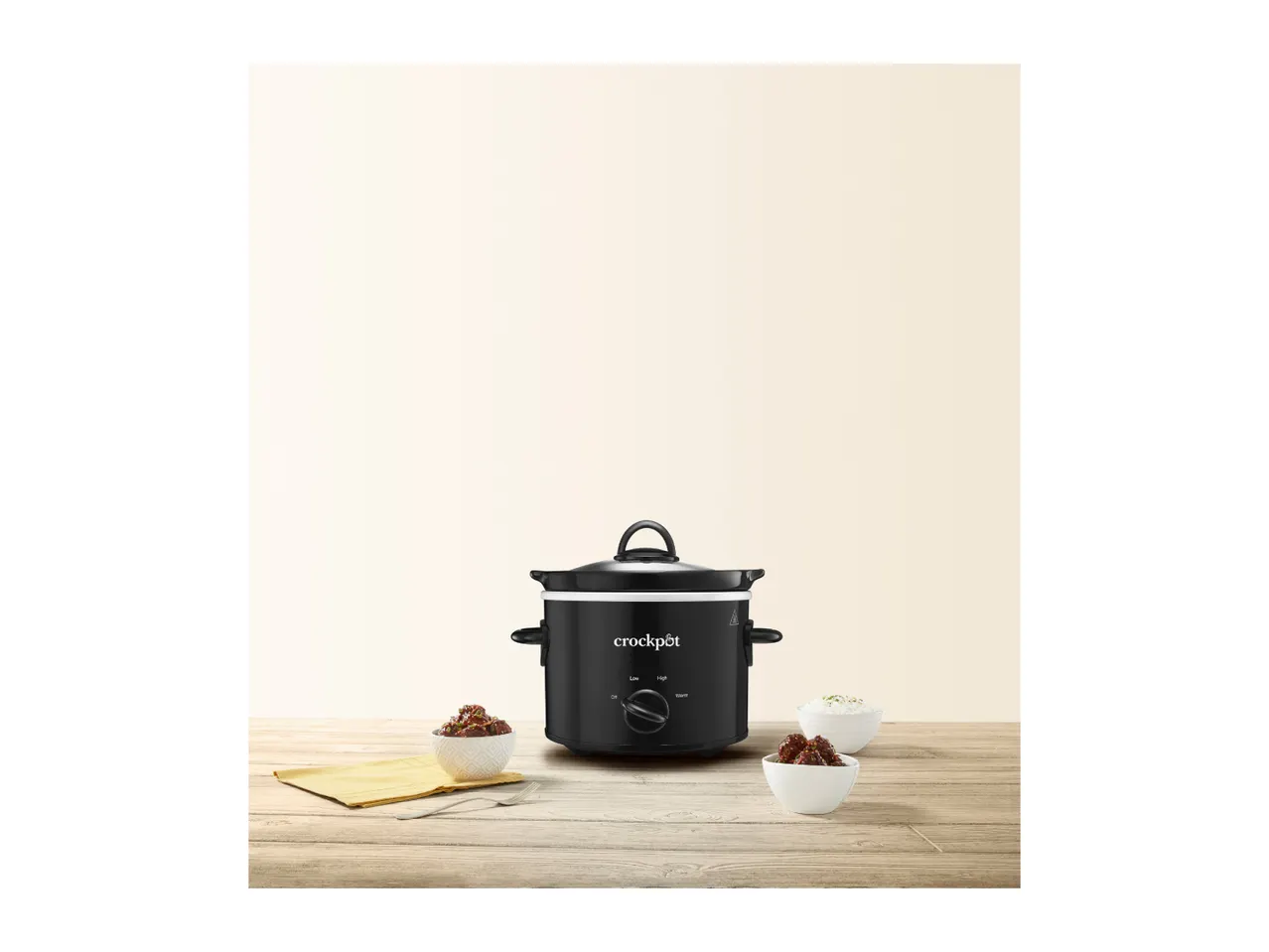 Black slow cooker on a wooden table with two bowls of food and a yellow napkin.
