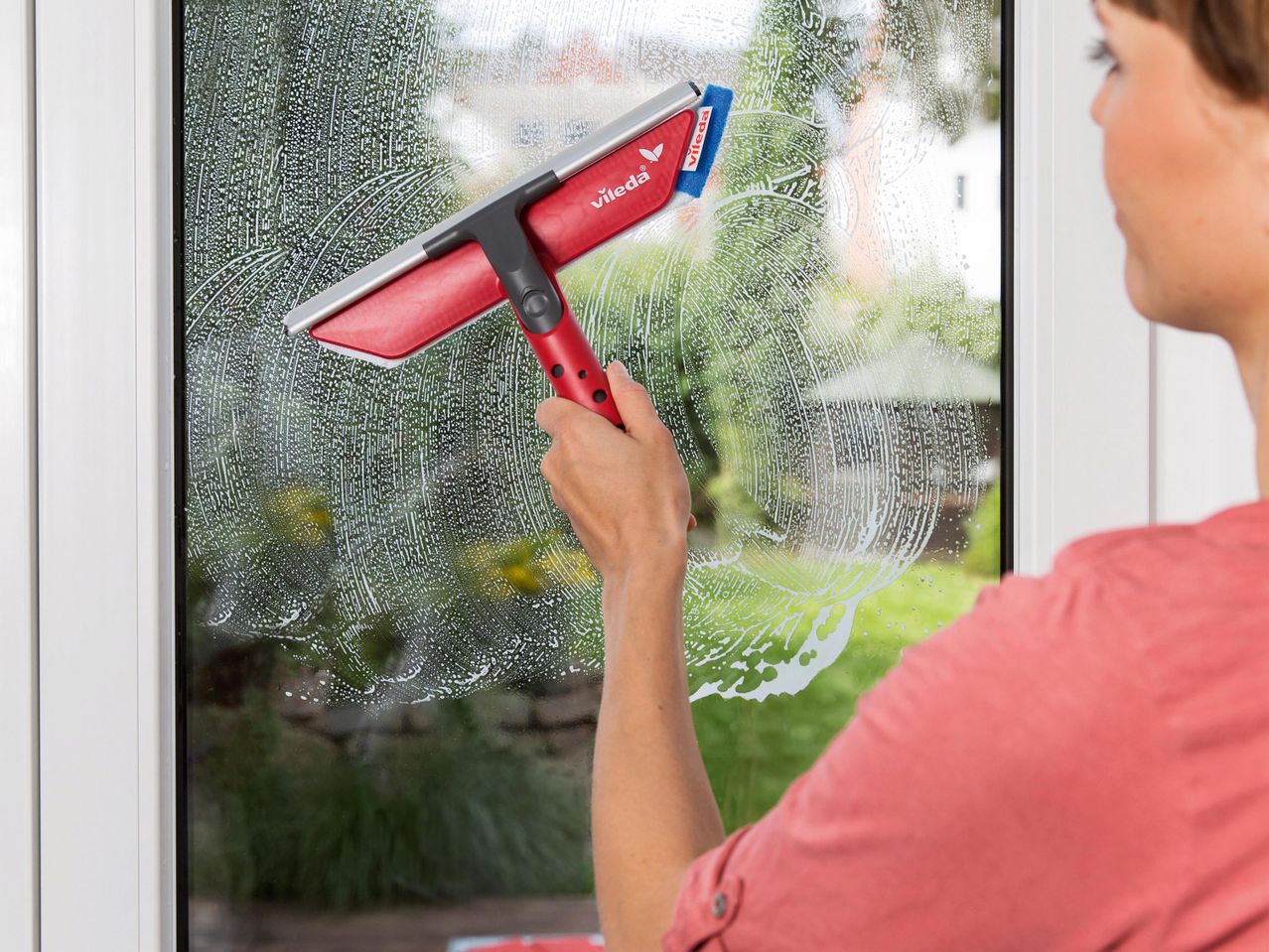 Nő ablaktisztítóval tisztítja az ablakot, szappanhabbal és zöld háttérrel.