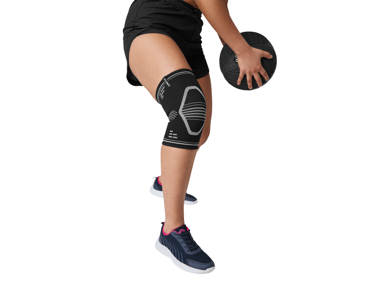 Person wearing a knee support and shorts, holding a medicine ball, with trainers visible.