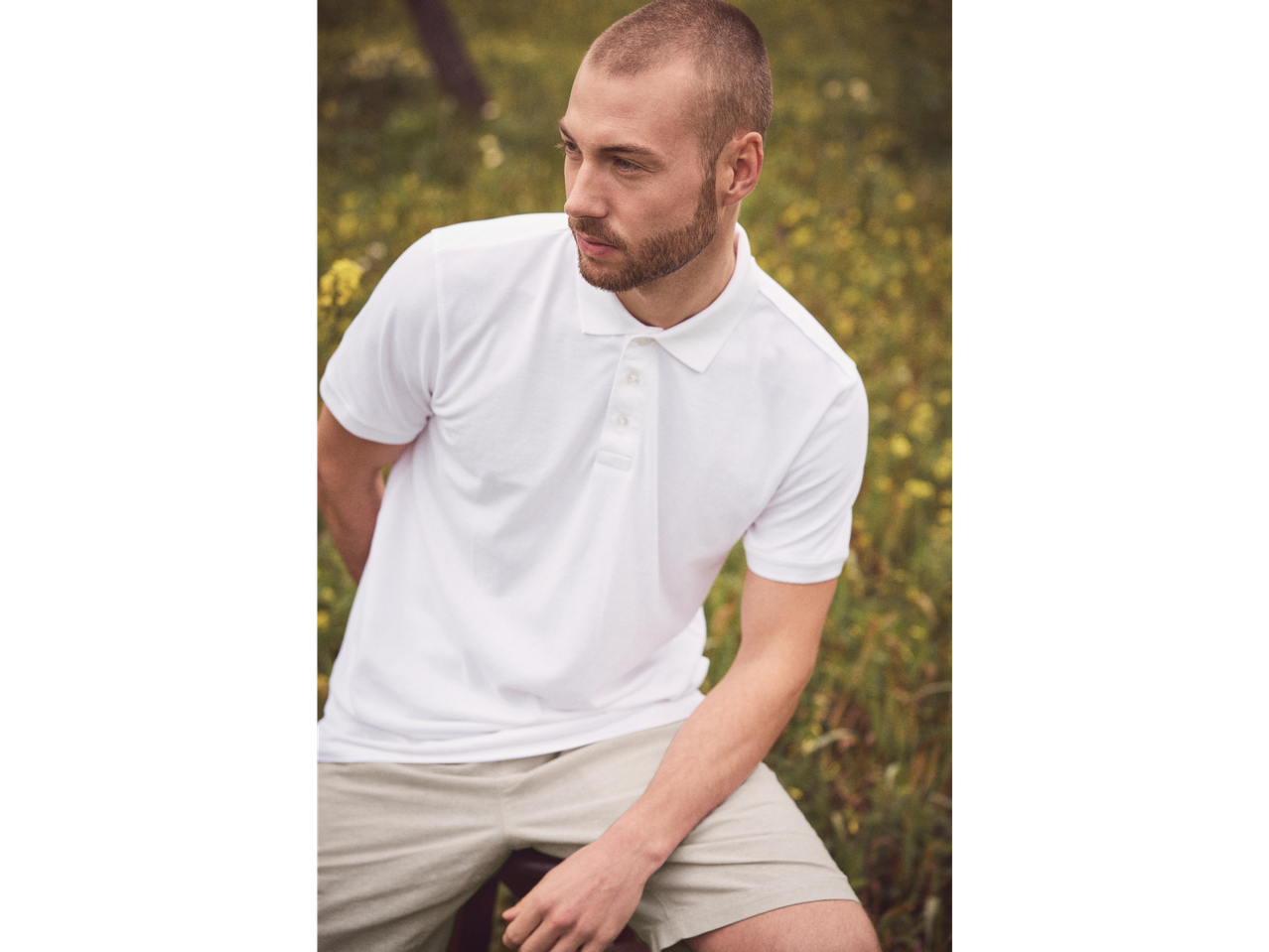 Model masculin cu polo alb și pantaloni scurți bej într-un câmp.