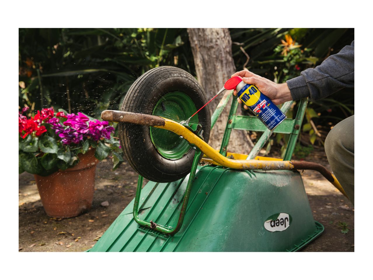 A person spraying lubricant on a wheelbarrow wheel, with potted flowers in the background.