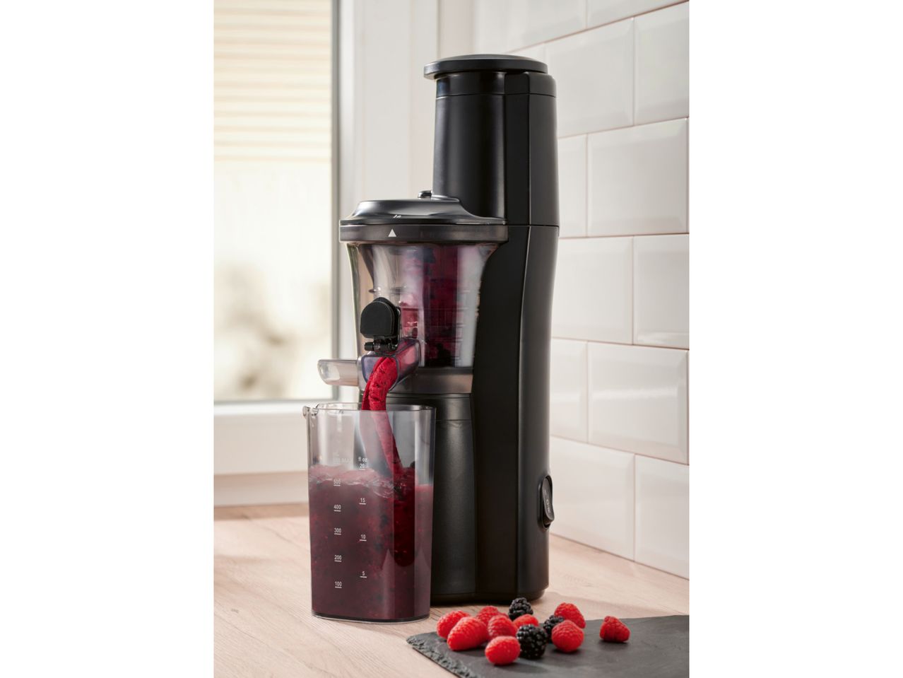 Black juicer extracting berry juice into a measuring cup on a wooden counter with fresh berries.