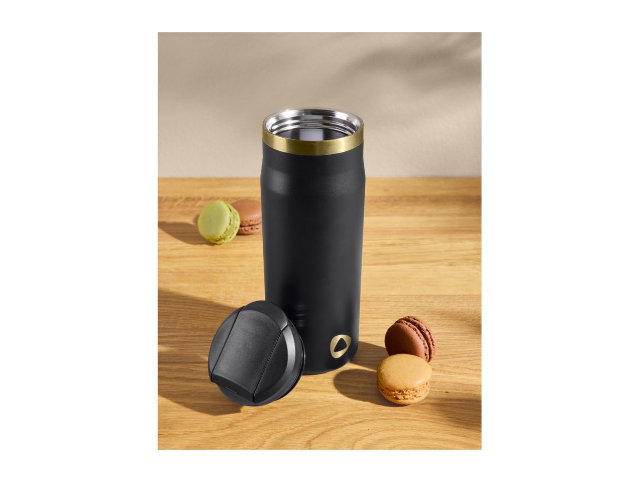 Black travel mug with a gold rim, next to its lid and three colorful macarons on a wooden surface.