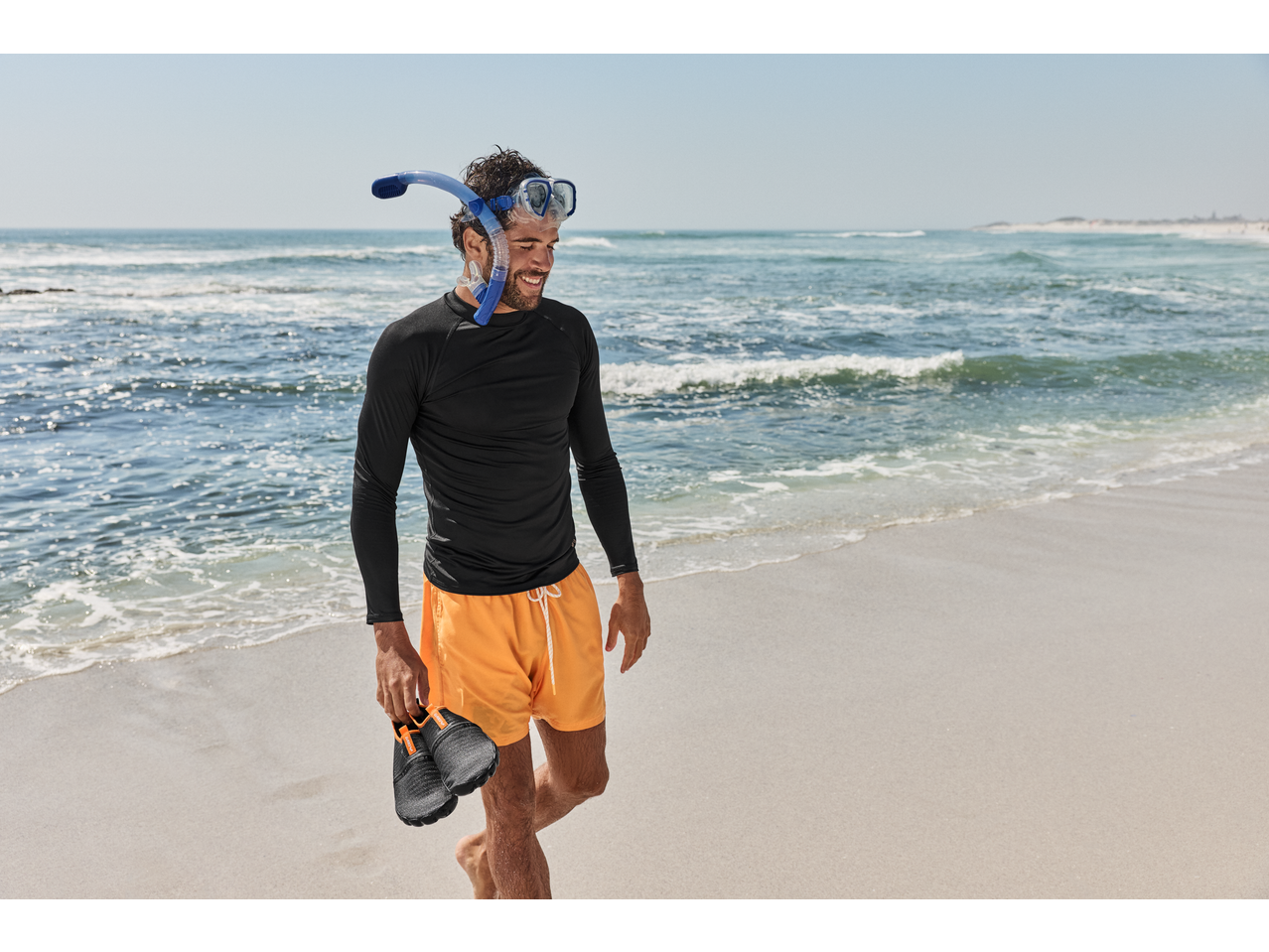 Man in CRIVIT aqua shoes, rash guard, and snorkel mask walking on a beach.