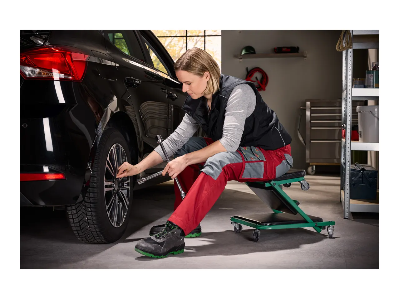 Woman using a creeper seat to change a car tire with a lug wrench in a garage.