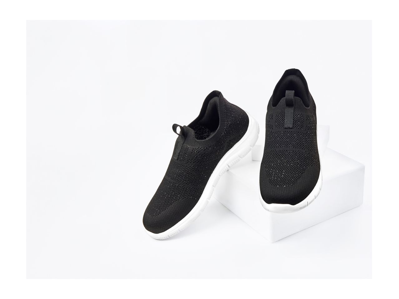 Pair of black slip-on sneakers with white soles, one standing on a white block.