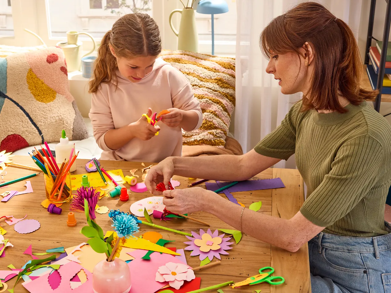 Майка и дъщеря правят цветя от хартия на дървена маса с цветни моливи и лепило.