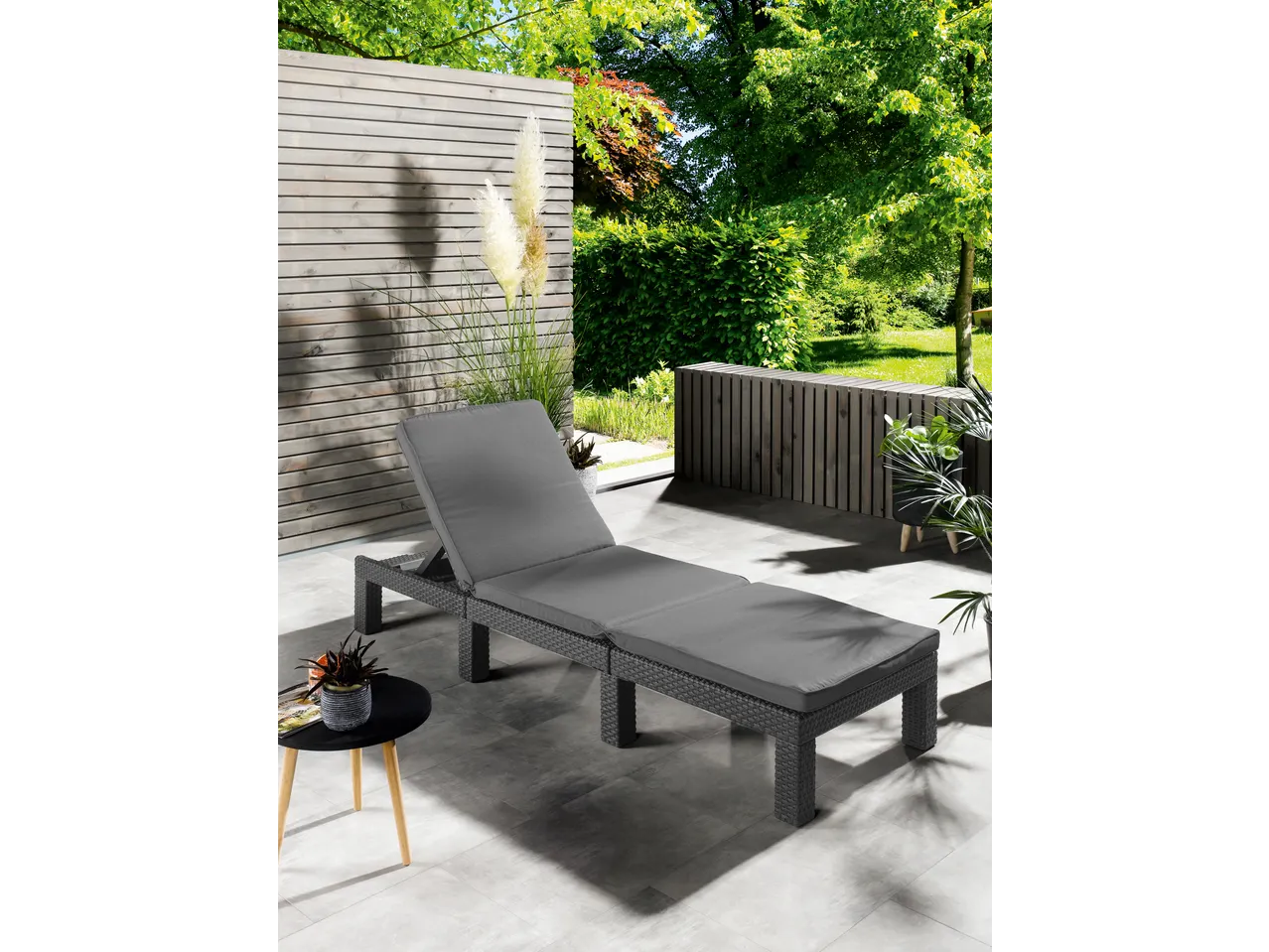 Outdoor lounge chair with grey cushions on a patio with wooden fencing and greenery.
