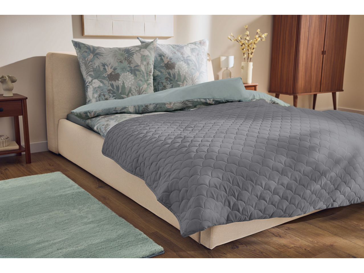 Quarto com cama com cabeceira de tecido, almofadas estampadas e edredão cinzento, com mesa de cabeceira e armário de madeira.