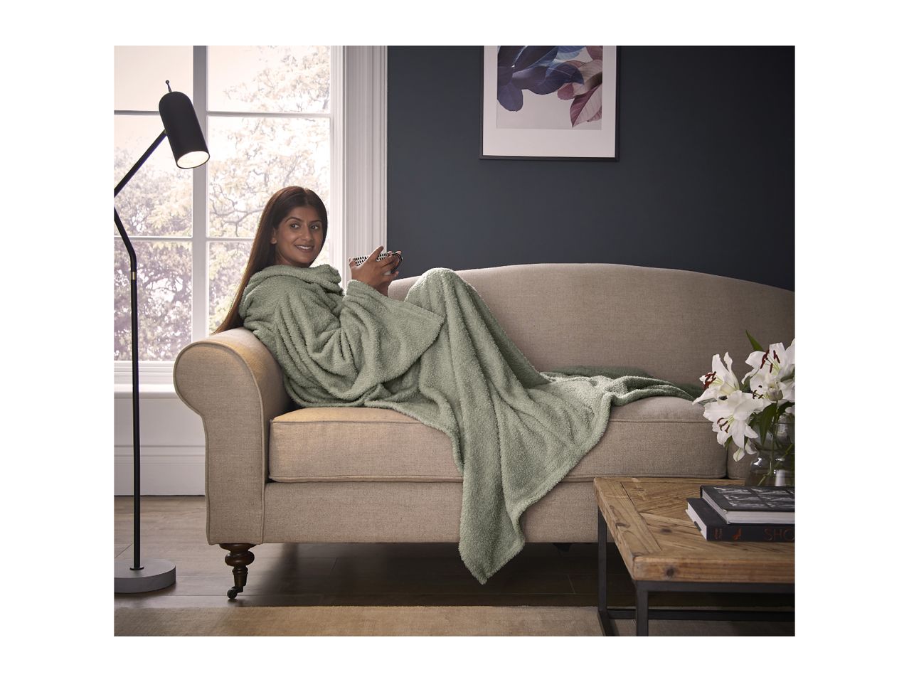A woman in a green hooded blanket on a sofa, holding a mug.