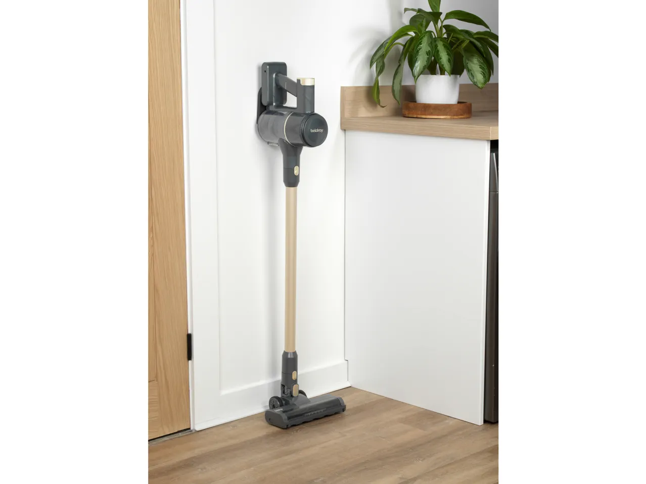 Cordless stick vacuum cleaner mounted on a white wall next to a wooden counter and plant.