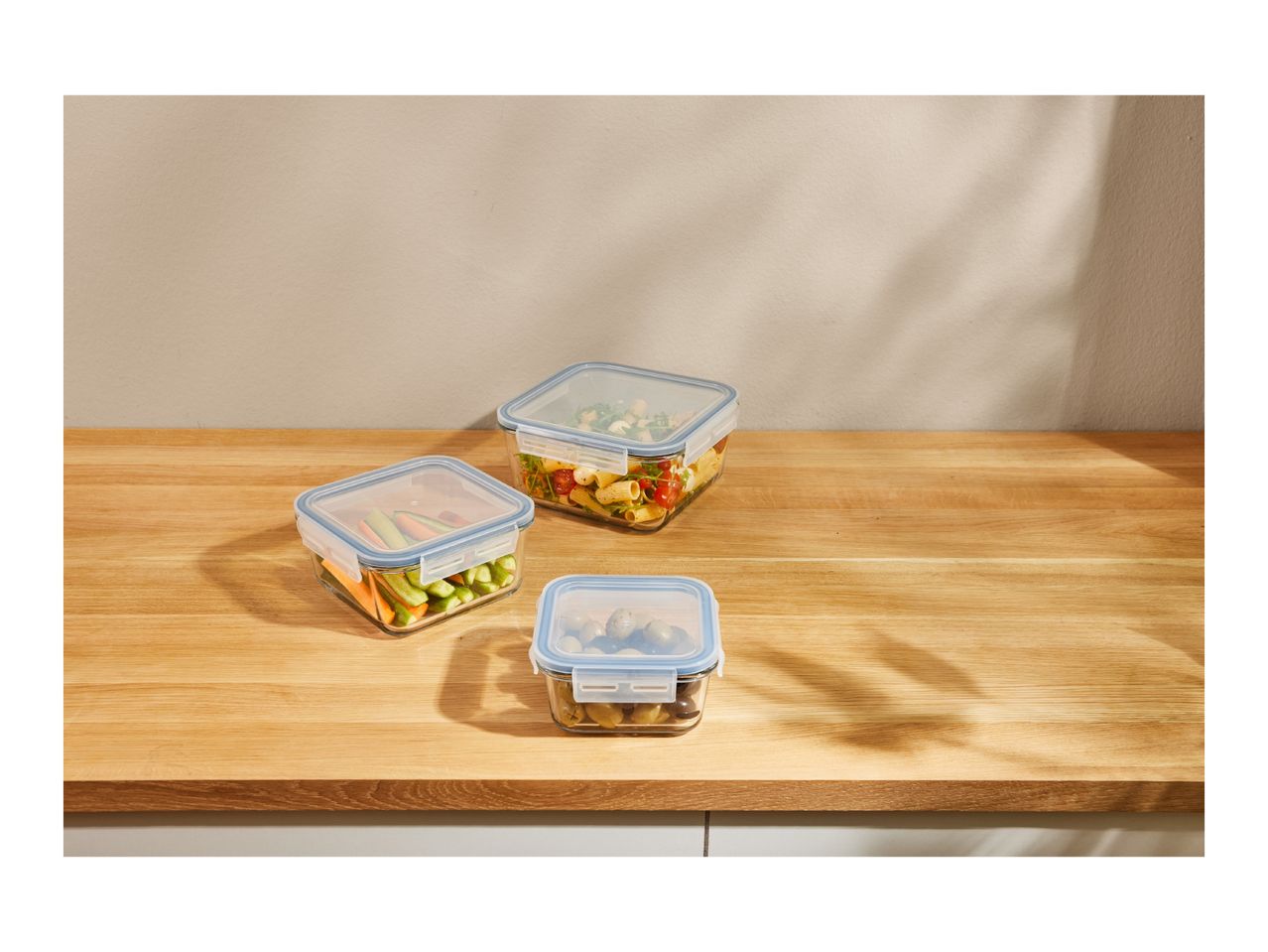 Three glass food storage containers with blue lids on a wooden counter.