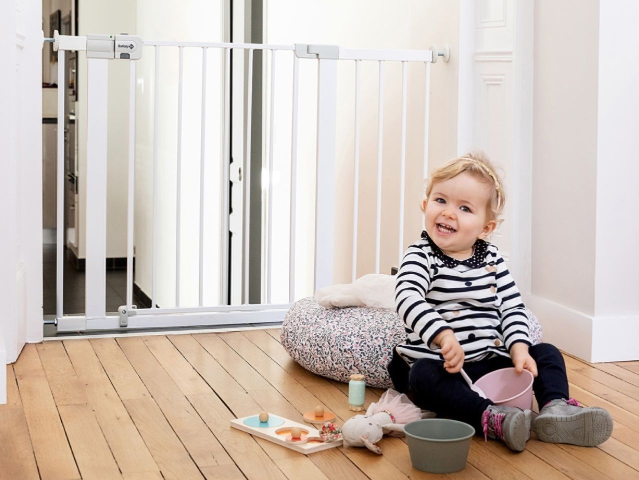 A baby sitting on the floor next to a safety gate, playing with toys and buckets.