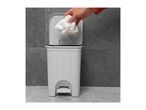 Hand dropping crumpled paper into a white woven pedal bin against a grey tiled background.