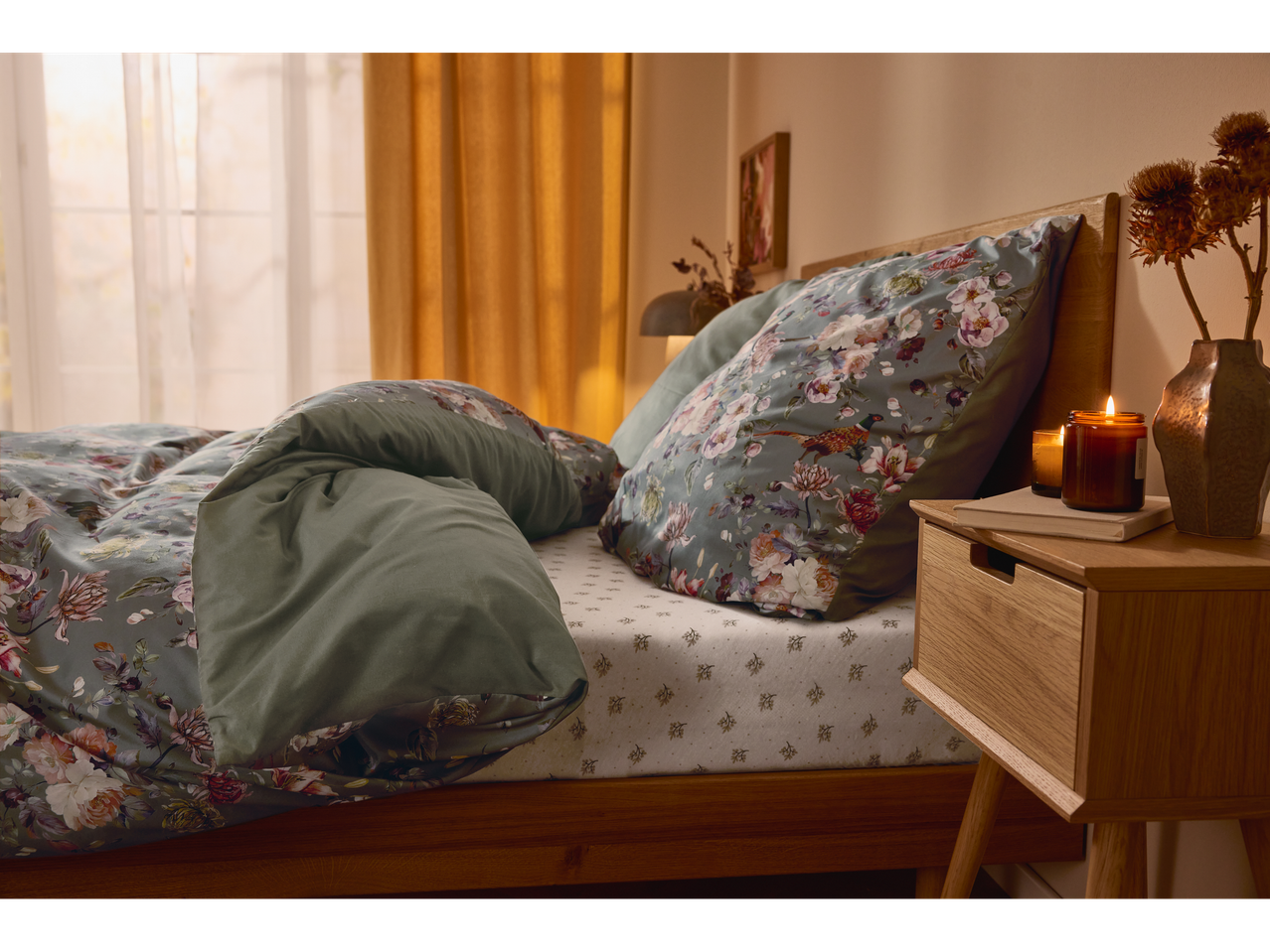 Un dormitor confortabil cu un pat din lemn, lenjerie de pat florală și o lumânare aprinsă pe noptieră.