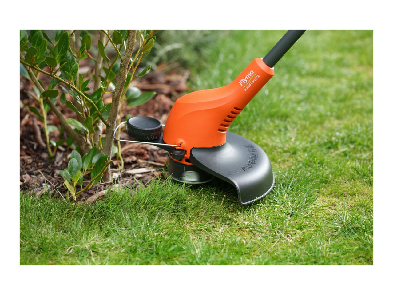 Orange grass trimmer cutting grass next to a green bush.
