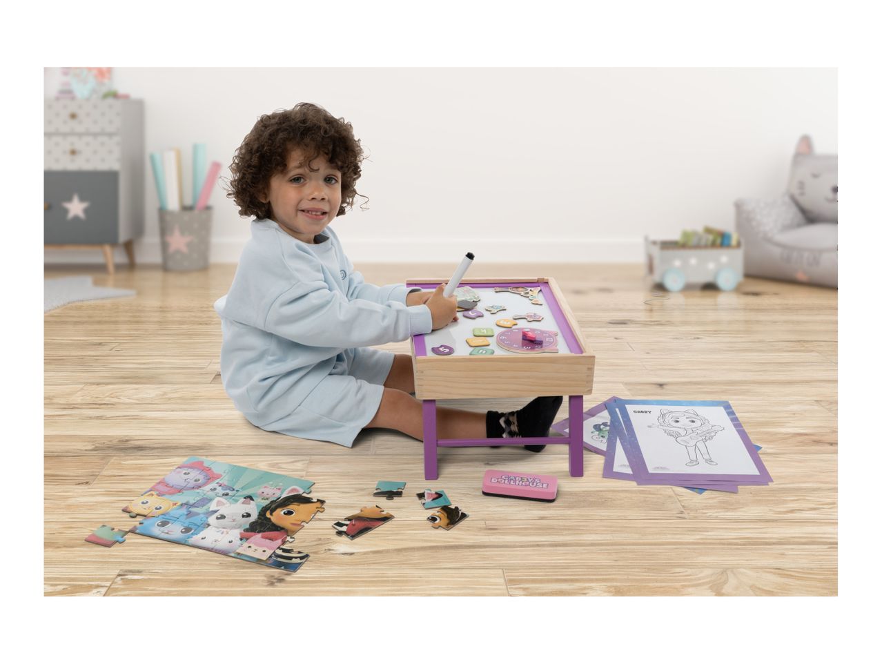 Child playing with a magnetic drawing board, puzzle, and coloring pages on a wooden floor.