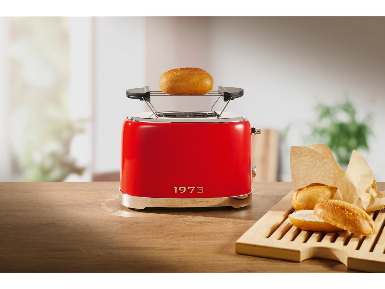 Red toaster with a bun on top, next to a wooden board with bread rolls.