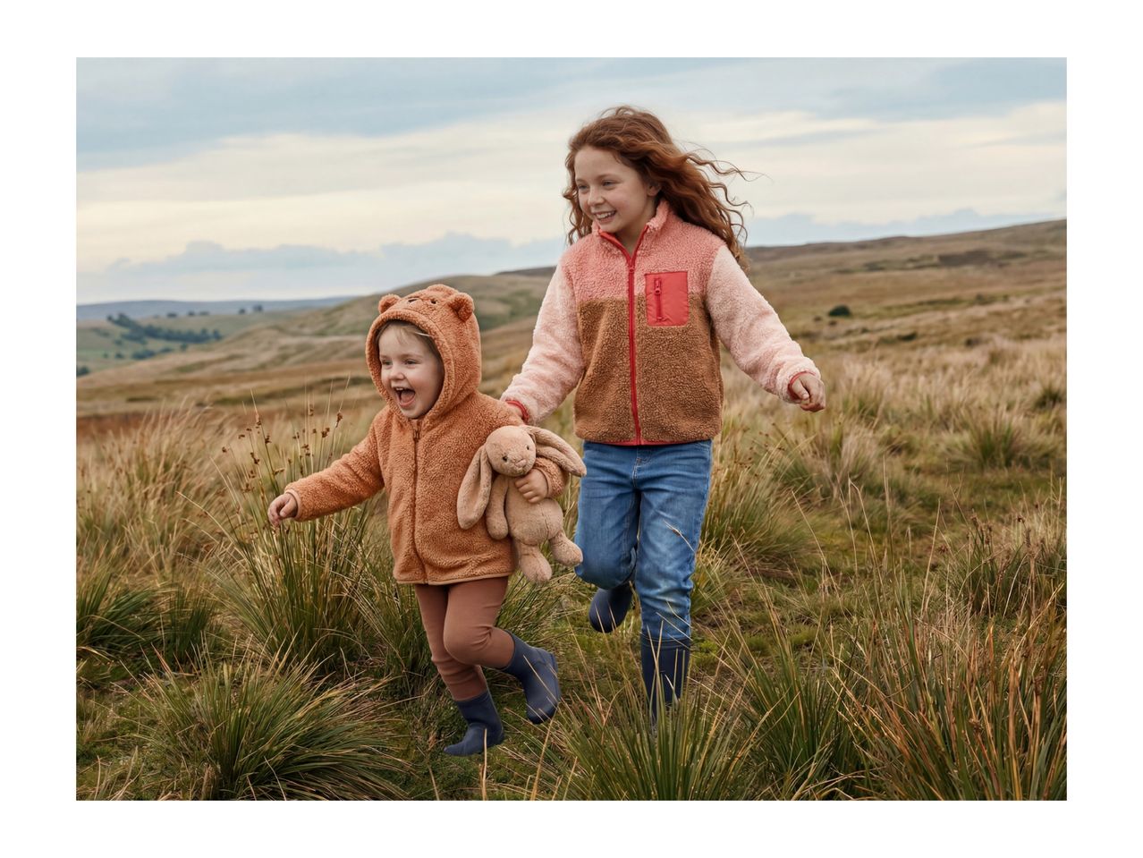Two happy children in fleece jackets and wellies running through tall grass.