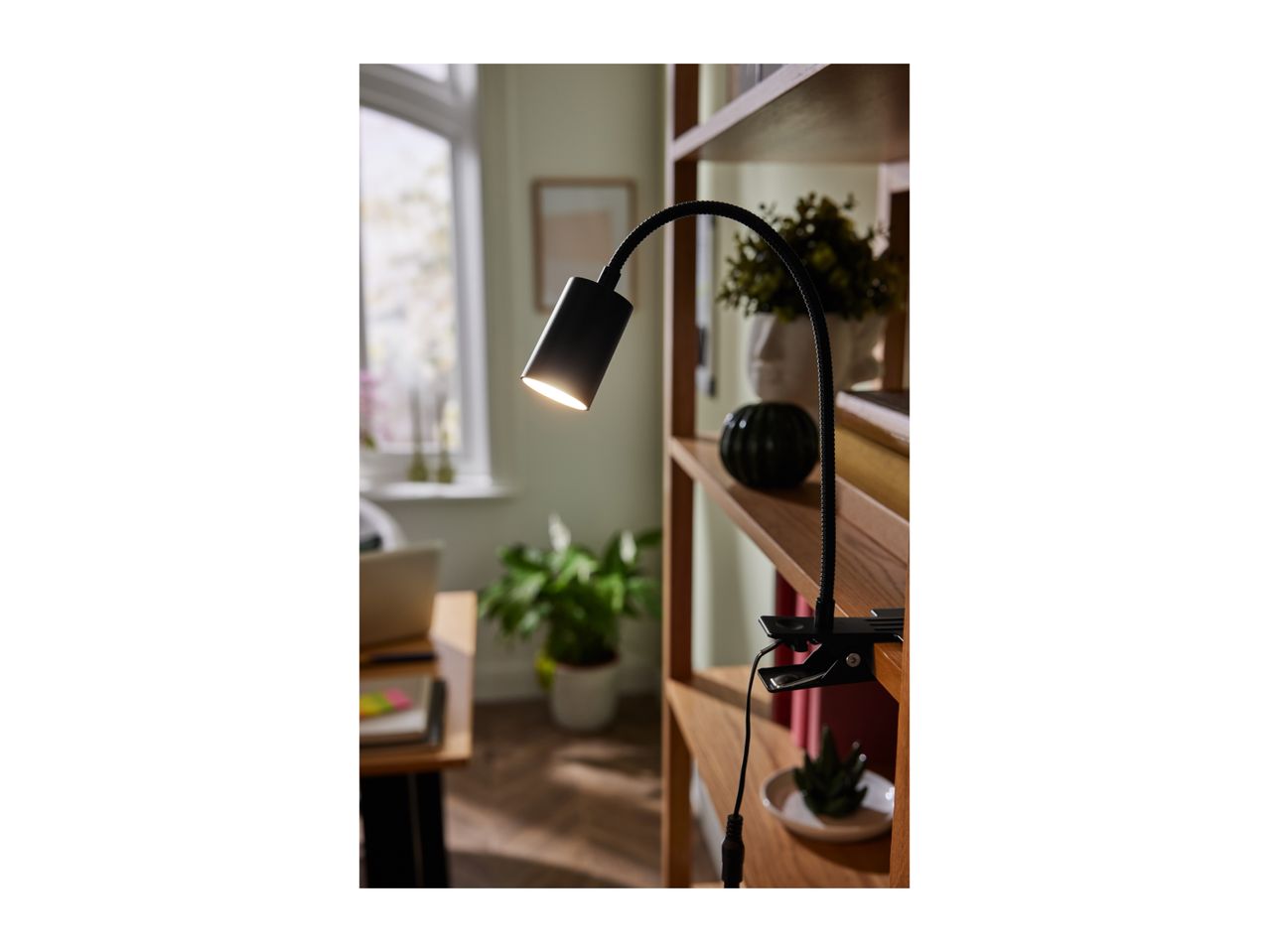 Black clip-on lamp illuminating a wooden bookshelf with plants and decor.