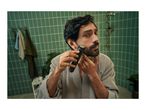Man in a bathrobe trimming his beard with an electric shaver in a bathroom with green tiles.