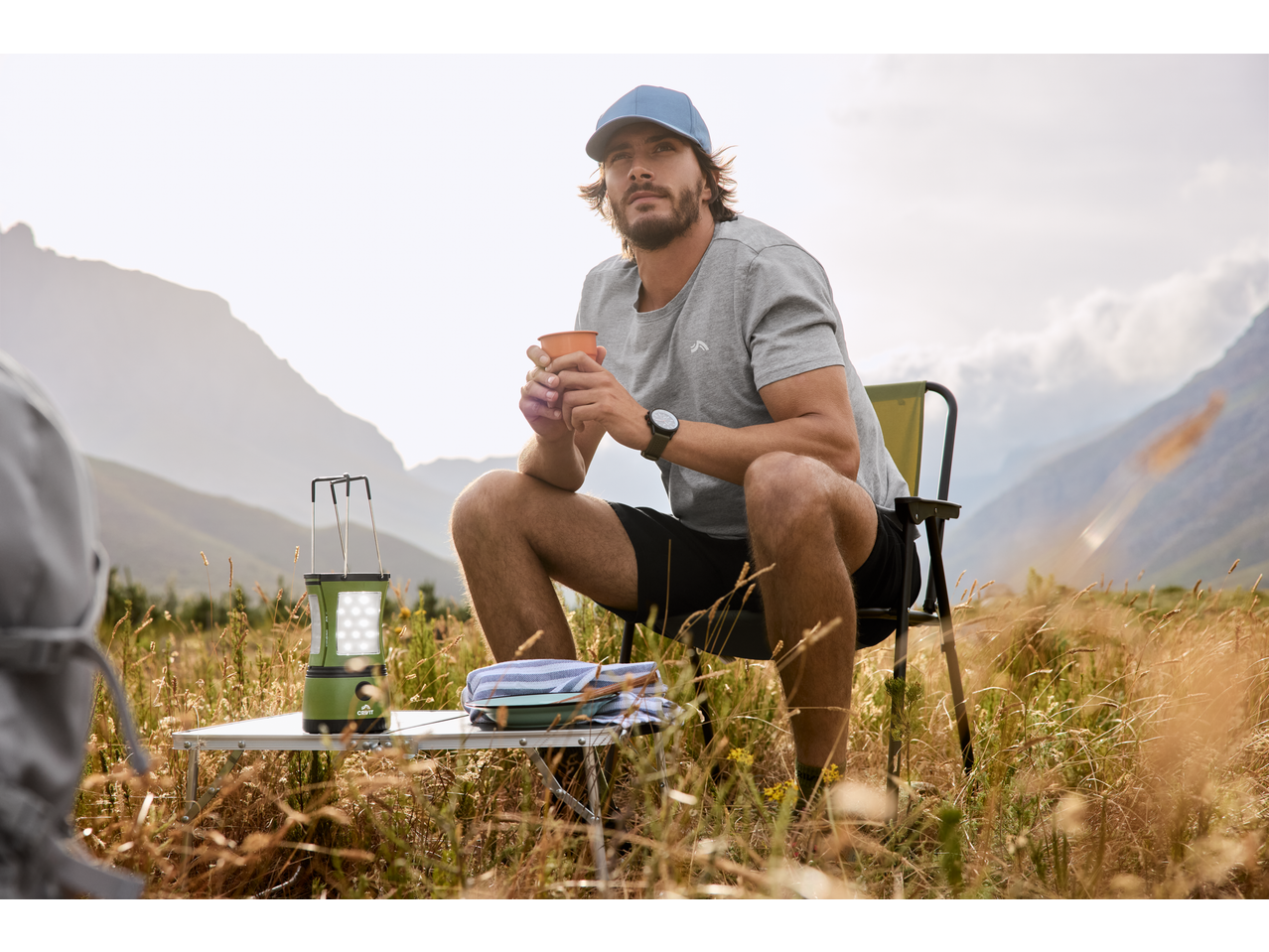 Man in cap and t-shirt sitting on a camping chair, holding a mug, with a lantern and table.