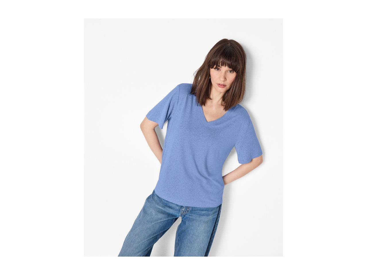 Woman in a blue V-neck t-shirt and jeans, leaning against a white wall.