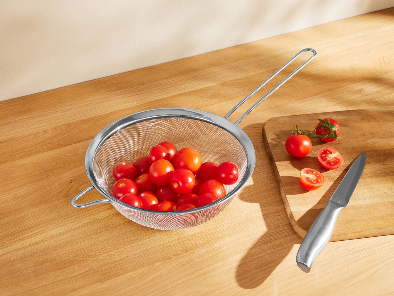 A stainless steel strainer filled with cherry tomatoes sits on a wooden counter next to a cutting board and knife.