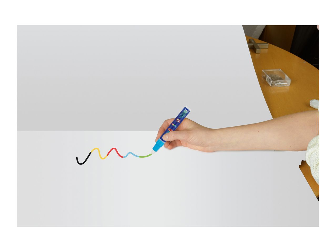 A hand drawing a colorful wavy line on white paper with a blue marker.