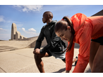 Two people in activewear jackets stretching outdoors, with a monument in the background.