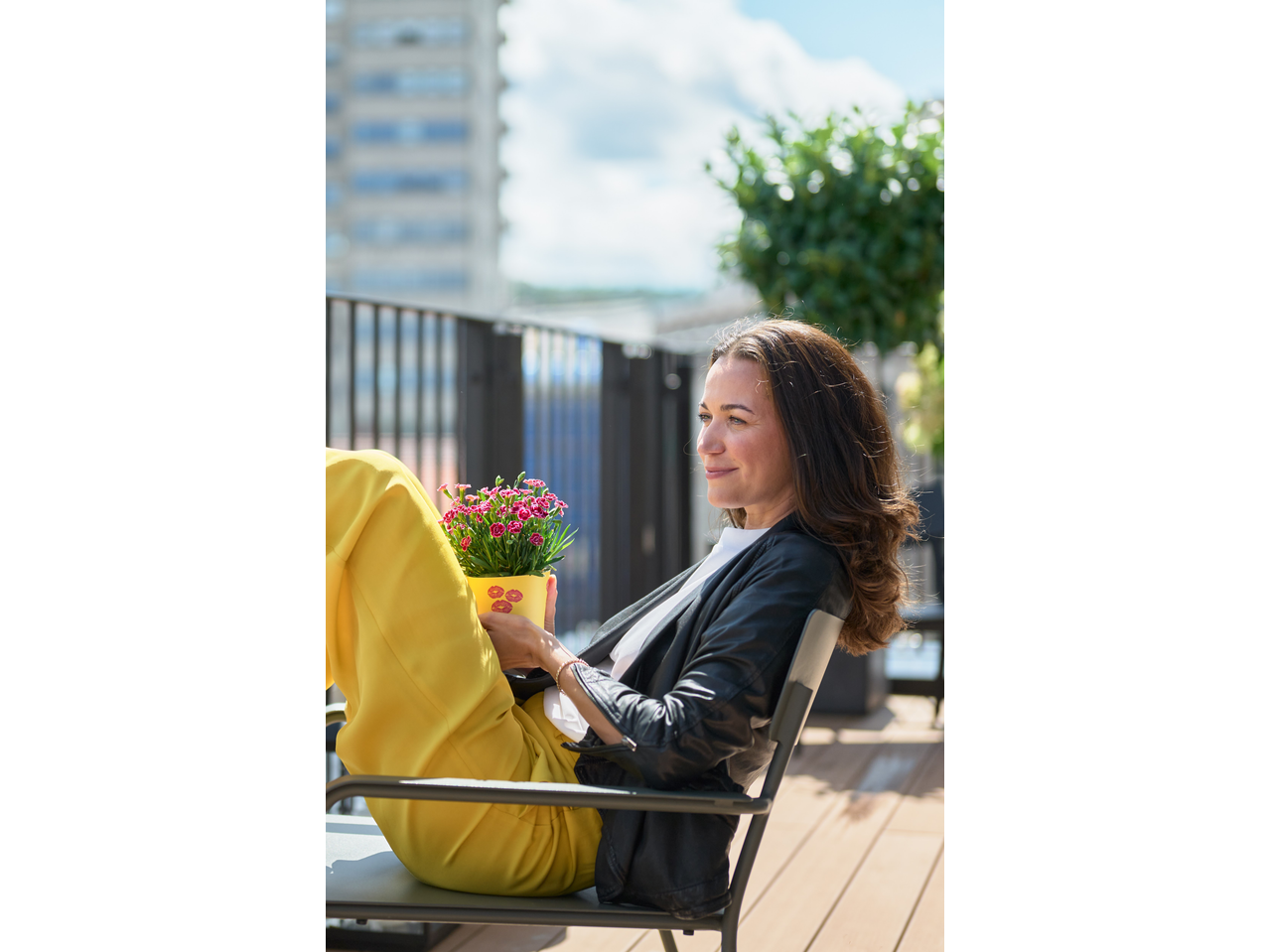 Woman in yellow pants and black jacket holding a pot of pink flowers with kiss marks.