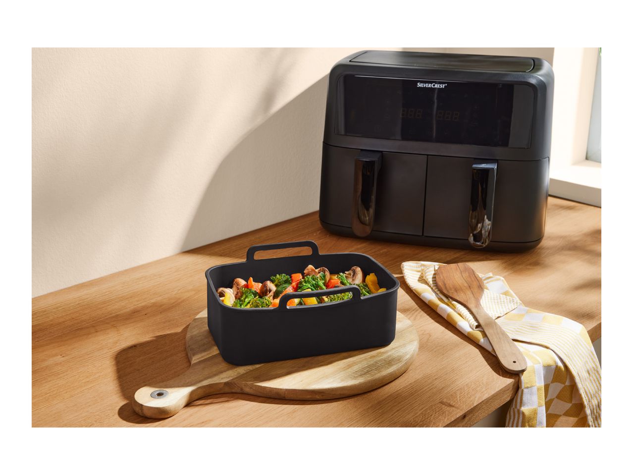 Dual air fryer with a black silicone baking dish filled with vegetables on a wooden counter.