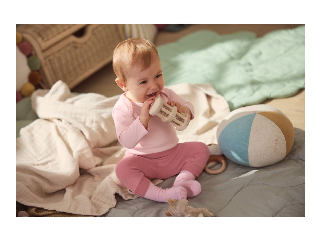 Happy baby in pink clothes playing with a wooden rattle and a soft ball.