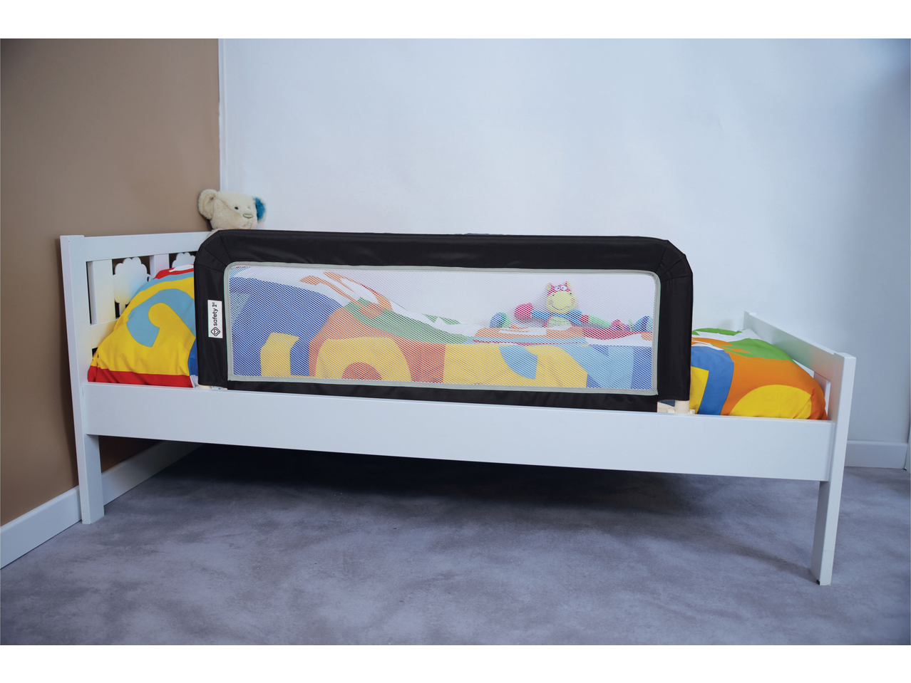 White toddler bed with a black bed rail, colorful bedding, and a teddy bear.