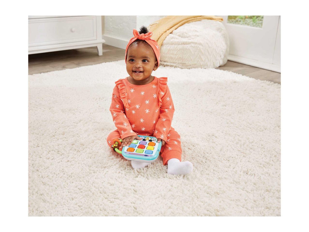Happy baby in a coral outfit with white snowflakes playing with a light-up learning toy on a white rug.
