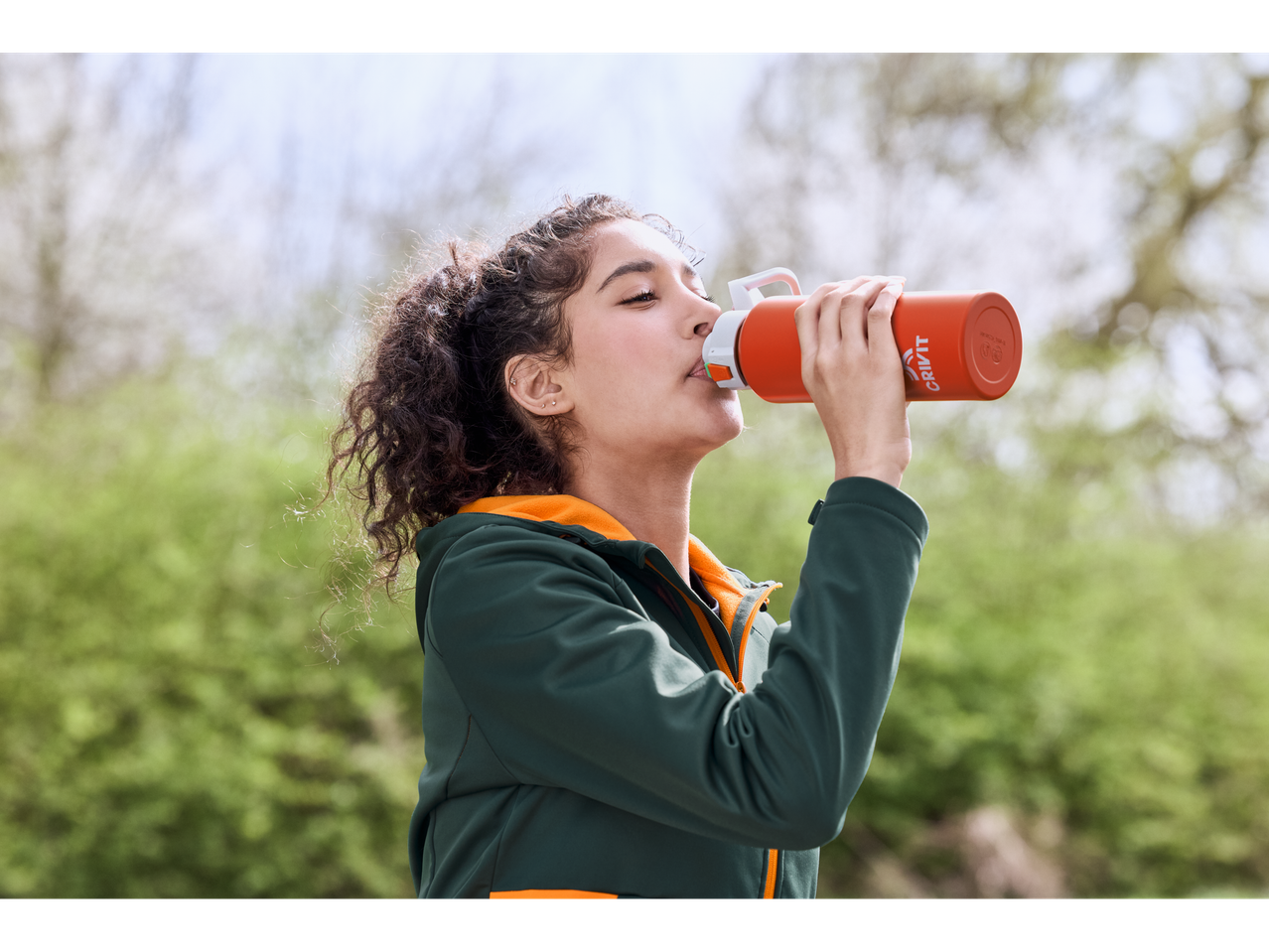 Woman drinking from an orange CRIVIT drink bottle outdoors.